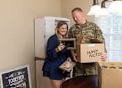 Thumbnail 12 of 21 - A man in military uniform is holding a box labeled "Family Photos" while a woman in a blue dress looks on.