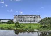Thumbnail 7 of 25 - A large white building with a grey roof is surrounded by greenery and water.