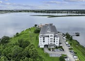 Thumbnail 6 of 25 - A large white building sits on a grassy area next to a body of water.