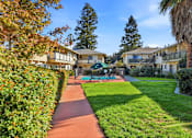 Thumbnail 3 of 33 - A pathway leads through a grassy area with a tennis court and apartment buildings in the background.