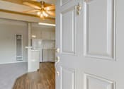 Thumbnail 14 of 32 - A white door with a gold handle is open to a room with a ceiling fan and a kitchen in the background.