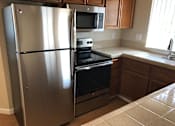 Thumbnail 12 of 26 - A stainless steel refrigerator stands in a kitchen with wooden cabinets and a black oven.