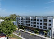 Thumbnail 20 of 21 - A large white building with balconies is surrounded by trees and has a parking lot in front.