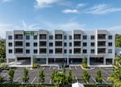 Thumbnail 12 of 21 - A large white building with a green sign on the top left corner and a parking lot in front.
