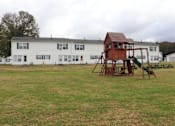 Thumbnail 10 of 12 - A playground with a wooden swing set in front of a white building.