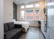 Thumbnail 5 of 20 - A grey couch sits in a living room with a window overlooking a red brick building.
