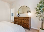 Thumbnail 12 of 42 - A bedroom with a white bed, a wooden chest of drawers, a round mirror, and a potted plant.