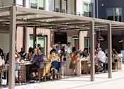 Thumbnail 38 of 63 - People are sitting at tables under a canopy outside a building.