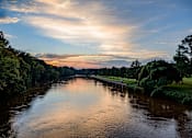 Thumbnail 19 of 21 - A serene river flows through a lush green landscape under a sky painted with hues of sunrise or sunset.