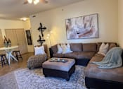 Thumbnail 11 of 21 - A living room with a brown couch, a grey ottoman, and a painting on the wall.