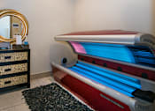 Thumbnail 19 of 21 - A tanning bed with a red and blue light is in a room with a black and gold dresser and a mirror.