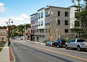 Thumbnail 2 of 14 - A street with cars parked on the side and buildings in the background.
