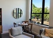 Thumbnail 5 of 12 - A living room with a grey couch, a wooden bookshelf, a round mirror, and a window with a view of trees.