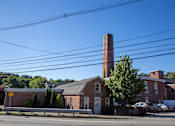 Thumbnail 7 of 12 - A brick building with a tall chimney is surrounded by trees and parked cars.