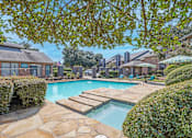Thumbnail 1 of 23 - A swimming pool surrounded by a stone patio and green bushes At The Pearl apartments in Shreveport LA