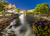 Thumbnail 24 of 81 - A river flows through a city at night with a hanging basket of flowers in the foreground.