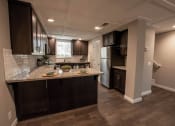 Thumbnail 16 of 27 - View from dining room into kitchen and partial view of stairwell to upstairs. Featuring stone counters, tile backsplash, brown cabinets, stainless appliances and wood look flooring