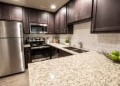 Thumbnail 3 of 26 - View of kitchen with stone counters, inlaid sink, stainless appliances, tile backsplash, brown cabinets, and wood look flooring.
