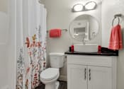 Thumbnail 19 of 36 - A white toilet with a floral shower curtain and a pink towel hangs on a wall next to a round mirror at Urban WalkApartments on 6th Avenue, Arizona 85016