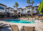 Thumbnail 17 of 20 - A swimming pool surrounded by lounge chairs and palm trees in front of a multi-story apartment building.