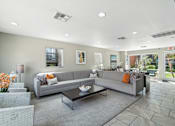 Thumbnail 15 of 26 - A grey couch with orange pillows sits in a living room with a glass coffee table in front of it.