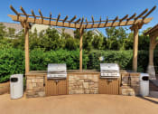 Thumbnail 22 of 54 - A large stone grilling station with two outdoor grills under a wooden pergola near trees