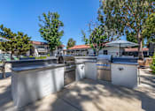 Thumbnail 4 of 28 - an outdoor kitchen with two grills and a table with chairs and umbrellas at Sage Creek Apartment Homes, Simi Valley California