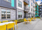 Thumbnail 1 of 18 - A patio with yellow chairs and tables is surrounded by apartment buildings.