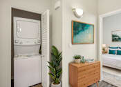 Thumbnail 12 of 28 - A small room with a white fridge, a wooden dresser, a potted plant, and a painting on the wall.