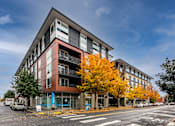 Thumbnail 1 of 25 - A modern building with a red and grey facade stands on a street corner with trees showing autumn colors.