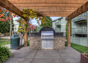 Thumbnail 1 of 16 - A stone and tile outdoor kitchen with a grill and sink under a wooden pergola.
