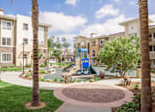 Thumbnail 17 of 21 - A playground with a blue slide is surrounded by palm trees and apartment buildings.