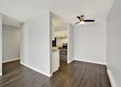 Thumbnail 24 of 28 - A room with a ceiling fan and a kitchen area in the background.