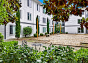 Thumbnail 13 of 28 - A white building with a gravel courtyard and a tree with a brown trunk.