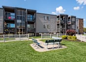 Thumbnail 12 of 15 - A modern apartment complex with a green lawn and benches in front.