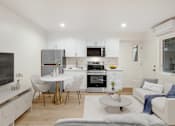 Thumbnail 18 of 45 - A modern kitchen with white cabinets and appliances, a dining table with chairs, and a living area with a couch.