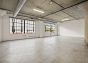 Thumbnail 14 of 92 - A large empty room with concrete floors and walls, two windows with bars, and exposed ceiling beams.