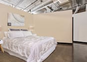 Thumbnail 16 of 123 - A bed with a white comforter and two pillows sits in a room with a concrete floor and a white wall with a painting and a lamp.