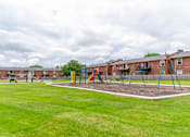 Thumbnail 7 of 15 - A playground with swings and a slide in front of a brick building.