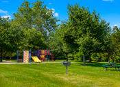 Thumbnail 3 of 16 - Picnic area at Cambridge Square Apartments in Greenwood, featuring a picnic table, a playground, and a BBQ grill.