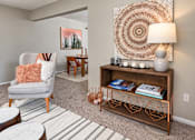 Thumbnail 26 of 29 - A living room with a grey armchair, a wooden coffee table, a wooden bookshelf, and a large woven wall hanging.
