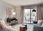 Thumbnail 24 of 29 - A living room with a grey sofa, a grey armchair, a wooden side table, a wooden coffee table, a lamp, a rug, and a large window with curtains.