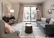Thumbnail 23 of 29 - A living room with a grey couch, a grey armchair, a wooden side table with a lamp, a round wooden table, a rug, and a large window with curtains.