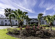 Thumbnail 19 of 19 - A white building with arched windows is surrounded by a well-manicured garden with palm trees.