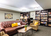 Thumbnail 15 of 26 - A living room with a brown leather couch, a chair, a coffee table, and a bookshelf filled with books.