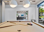 Thumbnail 15 of 53 - A pool table with a cue stick and balls is in the foreground of a room with a view of a dining area and outdoor patio.