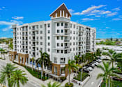 Thumbnail 2 of 40 - A large white building with a brown roof and balconies is surrounded by palm trees.