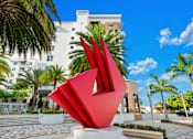 Thumbnail 29 of 40 - A large red sculpture in the foreground with a white building and palm trees in the background.