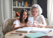 Thumbnail 28 of 28 - a young girl and older woman sitting at a table together looking at a cell phone