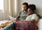 Thumbnail 6 of 8 - a man and a woman laying in bed looking at a laptop with natural light streaming in the window